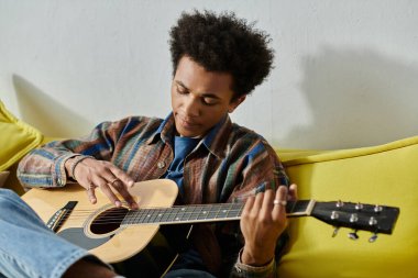 A young African American man sitting on a couch, playing an acoustic guitar while talking on the phone. clipart