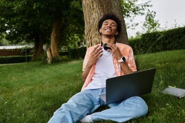 Young african american man sitting on grass, using laptop.