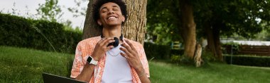 Young African American man sitting on tree in park with laptop in lap.