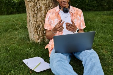 A young man, laptop open, sits serenely under a tree in a park.