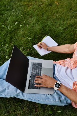 Young African American man sits on grass with laptop in a serene park setting.