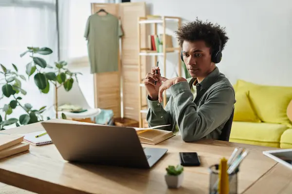 stock image A young African American man studying online at home, sitting at a desk with headphones and a laptop.