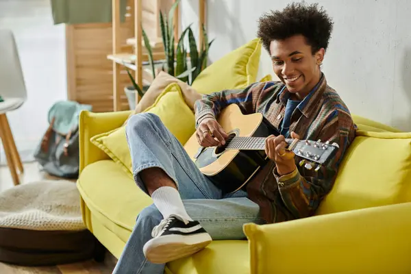 stock image A young man strums an acoustic guitar while seated on a vibrant yellow couch.