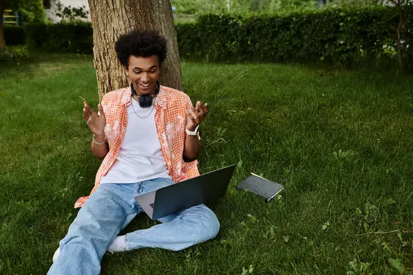 stock image Young African American man sitting on grass, working on laptop in the park.