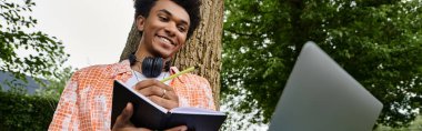 A young African American man holding a laptop in front of a tree in a park.