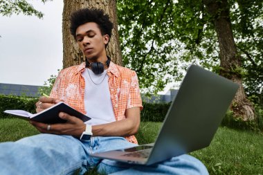 Young man immersed in work with laptop and headphones under tree.