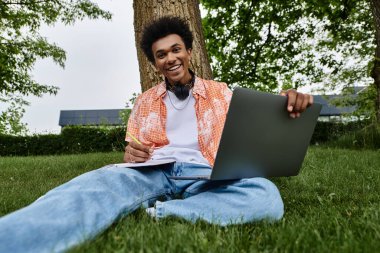 A young African American man sitting on grass with a laptop in a park.