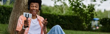 Young African American man sits under a tree, engrossed in his cell phone.