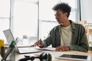 A young man, African American, focused and productive, works diligently on his laptop in a contemporary office space.