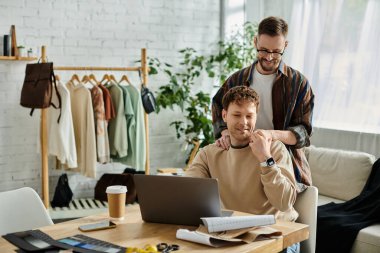 Bir adam dizüstü bilgisayarın önünde oturmuş, tasarımcı atölyesinde moda kıyafetler tasarlıyor..