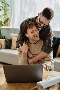 A man embraces his partner as the latter works on a laptop in a trendy designer workshop. clipart