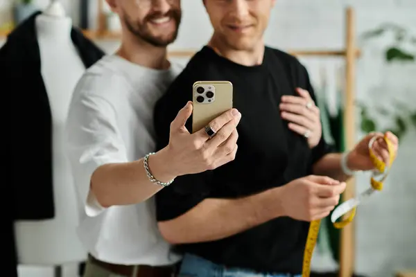 Stock image Two men, a gay couple, stand side by side with smartphone, working on trendy attire in their designer workshop.