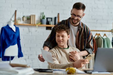 A man sits in a chair next to partner holding a piece for their collaborative fashion design project. clipart