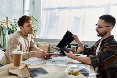 Tasarımcı atölyesinde çalışan iki adam. Kağıtlar ve dizüstü bilgisayarlar üzerinde çalışıyorlar..