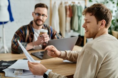 İki adam, bir masa ve bir dizüstü bilgisayarla birlikte moda tasarımları üzerinde çalışıyor..