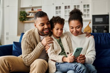 Afro-Amerikan aile kanepede kaynaşıyor, hepsi cep telefonu ekranına odaklanmış..