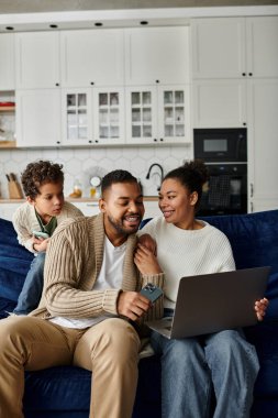Afro-Amerikan ailesi mavi bir koltukta otururken dizüstü bilgisayarla iş birliği yapıyor..