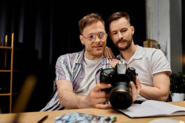 Two men, in casual outfits, sit at a table holding a camera. clipart