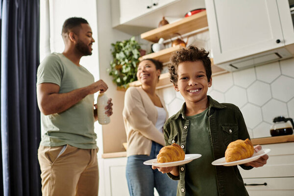 A young boy carefully holds two plates of food with his parents on backdrop.