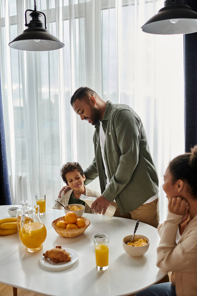 A man stands over a table heaped with a bounty of food, near son and wife.