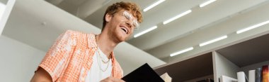 A man laughs while holding a book in a library. clipart