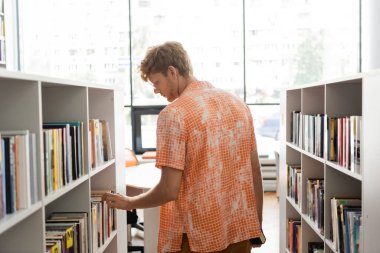 A young man stands before a bookshelf filled with books, immersed in study. clipart