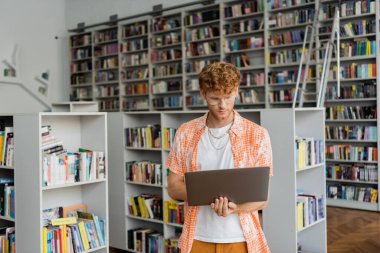 Man in front of books, holding laptop. clipart