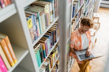 Genç bir adam kütüphanedeki kitapların arasında dizüstü bilgisayarına odaklanmış..