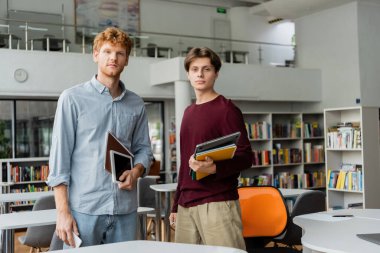 İki genç adam kütüphanede duruyor, her biri bir kitap tutuyor..