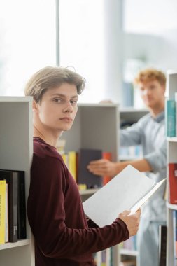 Arkaplanda arkadaşıyla birlikte kitaplığın önünde kitap tutan bir adam..