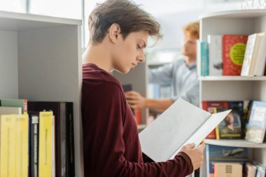 Kitaplara gömülmüş genç bir adam, raflarla çevrili, kütüphanede, arkaplanda arkadaşıyla.