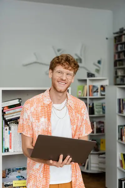 stock image Young man stands at bookshelf with tablet in hand.