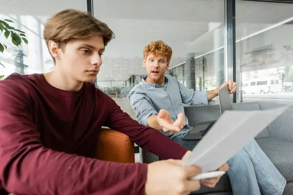 Stock image A man deep in thought while holding a paper on a couch, next to his friend.