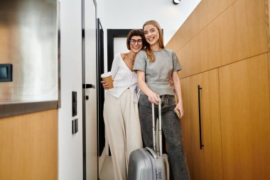 A young lesbian couple stands in a hotel room, ready to embark on a journey, holding coffee cups and luggage. clipart