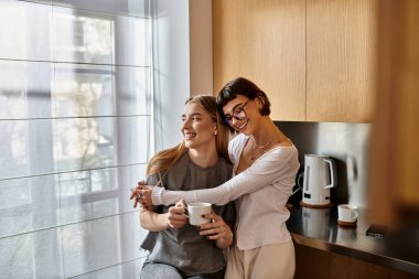 İki genç kadın, şık bir lezbiyen çift, modern bir otel mutfağında yan yana duruyorlar, ellerinde kahve fincanları var..