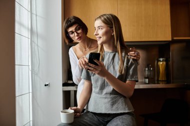 Genç lezbiyen çift kahveden zevk alıyorlar, tezgahın üzerinde oturuyorlar, ve bir otel odasının mutfağında cep telefonuna sarılıyorlar..