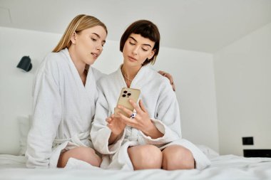 Banyo cüppeli iki kadın bir yatakta oturuyor, cep telefonuna dalmış, bir otel odasında özel bir anı paylaşıyorlar..