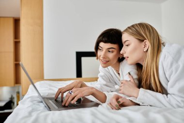 Yatakta dinlenen, rahat bir otel odasında dizüstü bilgisayarın keyfini çıkaran banyo cüppeli güzel bir lezbiyen çift..