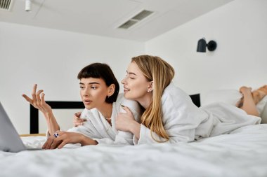 Banyo cüppeli iki kadın, dizüstü bilgisayara sarılmış bir yatakta dinleniyor..