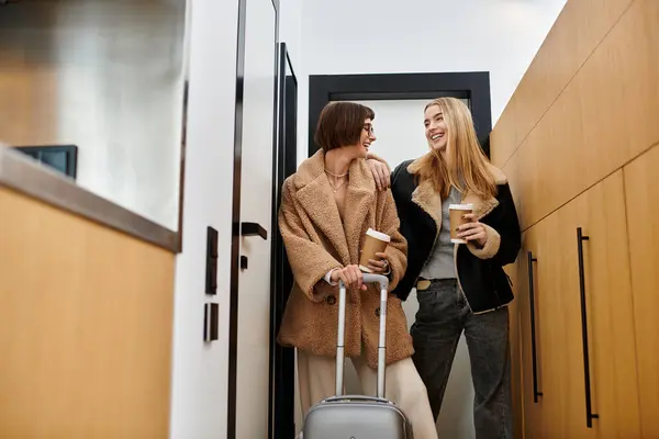 stock image A young lesbian couple stands side by side, exuding style and grace, with luggage in a hotel hall.