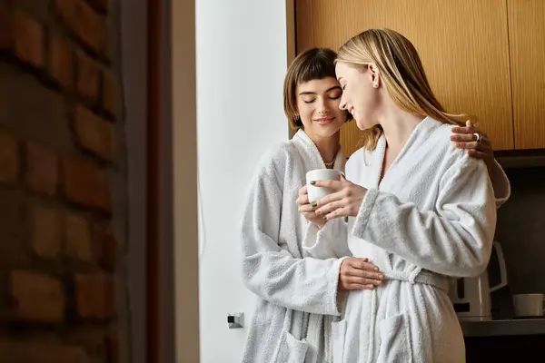 Stock image A young lesbian couple stands gracefully side by side in soft bathrobes inside a cozy hotel room.