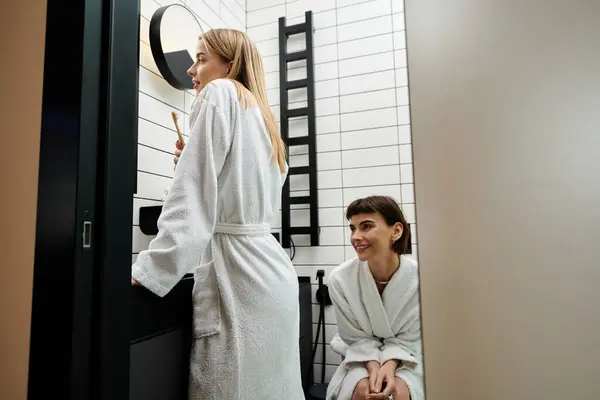 Stock image blonde woman in a bathrobe brushes her teeth while her partner watches in a hotel bathroom.