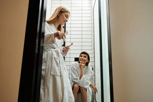 stock image A young lesbian couple in bathrobes, one sitting on a toilet and the other standing next to her inside a hotel bathroom.