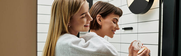 A young lesbian couple in bathrobes stand together in front of a mirror, gazing at their reflections with affection.