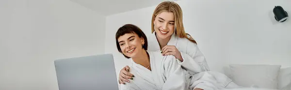 Stock image Young lesbian couple in white robes exploring something intriguing on a laptop together.