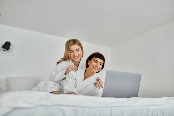 stock image Two young women in white robes, relaxing on a bed together and using a laptop.