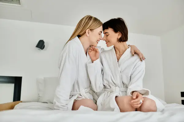 stock image A beautiful lesbian couple in white robes sit serenely on a bed inside a hotel room, exuding grace and peace.