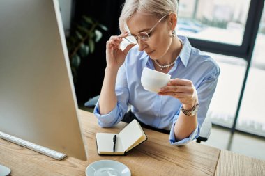 A middle aged businesswoman with short hair enjoys a cup of coffee while working in her office clipart