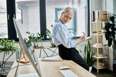 Kısa saçlı, odaklanmış orta yaşlı bir iş kadını masasında özenle çalışıyor, bir not panosuna notlar yazıyor.