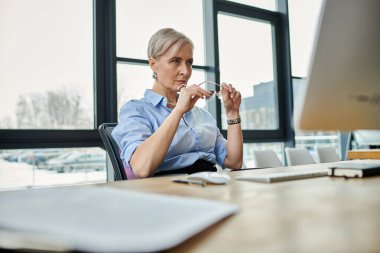 A middle-aged businesswoman with short hair works on a computer at a desk in a modern office clipart